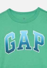 Πράσινο βαμβακερό t-shirt με στρογγυλή λαιμόκοψη, με μεγάλα γράμματα "GAP" σε μπλε διαβαθμίσεις στο μπροστινό μέρος. Λεπτή υφή και καθημερινό σχέδιο.
