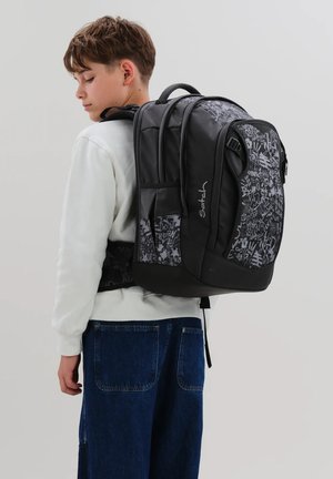 Teenager-Junge trägt ein weißes Sweatshirt und blaue Jeans und hat einen schwarz-grau gemusterten Satch-Rucksack mit gepolsterten Trägern auf dem Rücken.