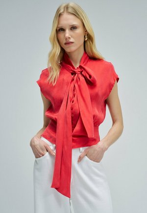 Mujer rubia con una blusa roja sin mangas con un gran lazo atado en el cuello, metida dentro de unos pantalones blancos de talle alto, con las manos en los bolsillos.