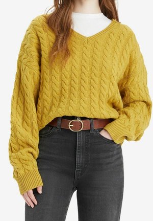 Femme portant un pull en maille torsadée jaune moutarde par-dessus une chemise blanche, un jean noir taille haute et une ceinture marron avec une boucle dorée.