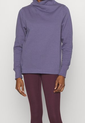 Person iført en langærmet lilla hættetrøje og mørkelilla leggings, stående med afslappede arme mod en ensfarvet baggrund.