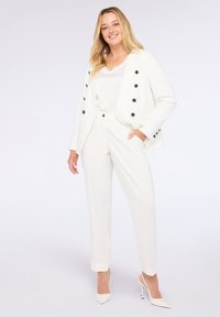 Bílý tailored oblek s blejzrem, který má černé knoflíkové detaily, v kombinaci s bílým topem a slim-fit kalhotami. Celý vzhled doplňují stiletto podpatky.
