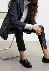 Personne assise sur une chaise portant un blazer sombre, un pull à col roulé blanc, un pantalon en cuir noir et des mocassins marron foncé.
