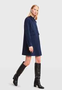 Námornícky modrá šaty s golierom a dlhými rukávmi, s predným gombíkovým zapínaním a bočnými vreckami. V kombinácii s čiernymi koženými čižmami do kolien. Hladký povrch.