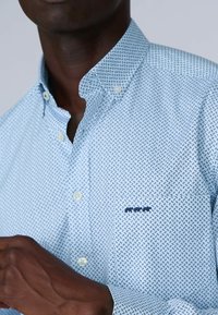 Chemise bleu clair à boutons avec un motif répétitif bleu marine, col button-down et une poche poitrine avec des détails de couture en marine.