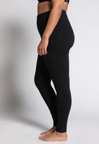 Leggings neri con vita elastica, texture liscia, design aderente e taglio alla caviglia, indossati da un modello di profilo.