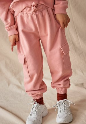 Pantaloni de trening roz cu o croială ajustată, talie elastică și buzunare laterale tip cargo, asortați cu adidași albi și șosete roșii cu țesătură ribbed.