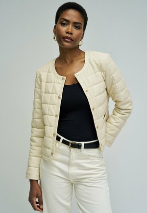 Femme portant une veste matelassée crème sur un top noir, pantalon blanc avec une ceinture noire, debout avec une main sur la hanche.