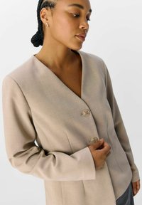 Beiger Blazer mit tiefem V-Ausschnitt, drei Knöpfen, taillierter Passform und strukturierten Schultern. Verfügt über eine glatte Stofftextur und dezente Nähte.