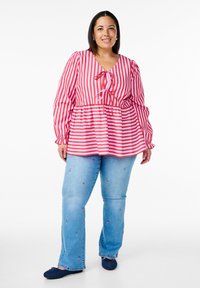 Sarkana un balta svītrota blūze ar kakla saiti, pūkainām piedurknēm un pepluma vidukli, kopā ar gaiši ziliem izšūtiem džinsiem un tumši ziliem apaviem.