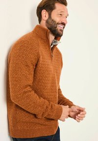 Rustfarbener Reißverschluss-Pullover mit einem strukturierten Fischgrätenmuster. Verfügt über einen hohen Kragen, gerippte Bündchen und Saum. Getragen über einem karierten Hemd.