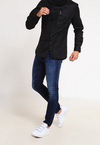Camisa negra de manga larga con un bolsillo en el pecho con cremallera, combinada con jeans ajustados azules y zapatillas blancas. Tejido suave y corte entallado.