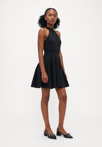 Robe noire sans manches avec un buste ajusté et une jupe évasée, présentant un décolleté haut et un tissu lisse. Associée à des chaussures à bride arrière noires.