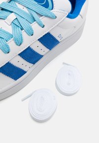Baskets blanches en cuir avec des accents en daim bleu et des lacets bleus. Présente trois rayures bleues et deux lacets blancs roulés à côté.