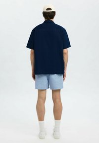 Kortärmad marinblå skjorta, ljusblå rutig shorts, vita sneakers och en beige keps, sedd bakifrån mot en ljus bakgrund.