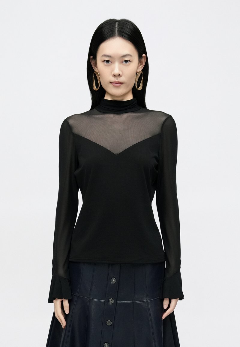 Fekete hosszú ujjú felső, áttetsző hálós nyakkal, V alakú kivágással, puha anyagból, fodros mandzsettákkal. Sötét, gombos szoknyával viselve.