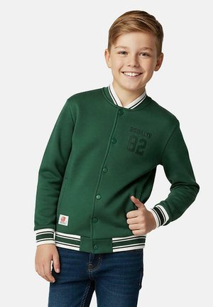 Jongen die lacht en duimen omhoog doet, draagt een groene jas met knopen en witte gestreepte manchetten en kraag, en blauwe spijkerbroek.