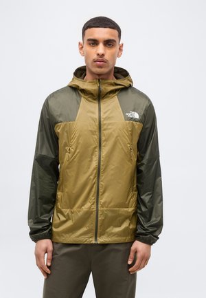 Άνδρας που φοράει καφέ και σκούρο πράσινο μπουφάν North Face με κουκούλα, εμπρός φερμουάρ και φερμουάρ σε τσέπες, κοιτάζοντας προς τα εμπρός σε απλό φόντο.