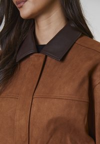 Kobieta w brązowej zamszowej kurtce z kontrastowym ciemnoskórzanym kołnierzem, pokazana od brody do klatki piersiowej.