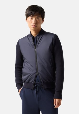 Young man wearing a navy bomber jacket with zippered front and knit sleeves, standing with one hand in pocket against a plain background.