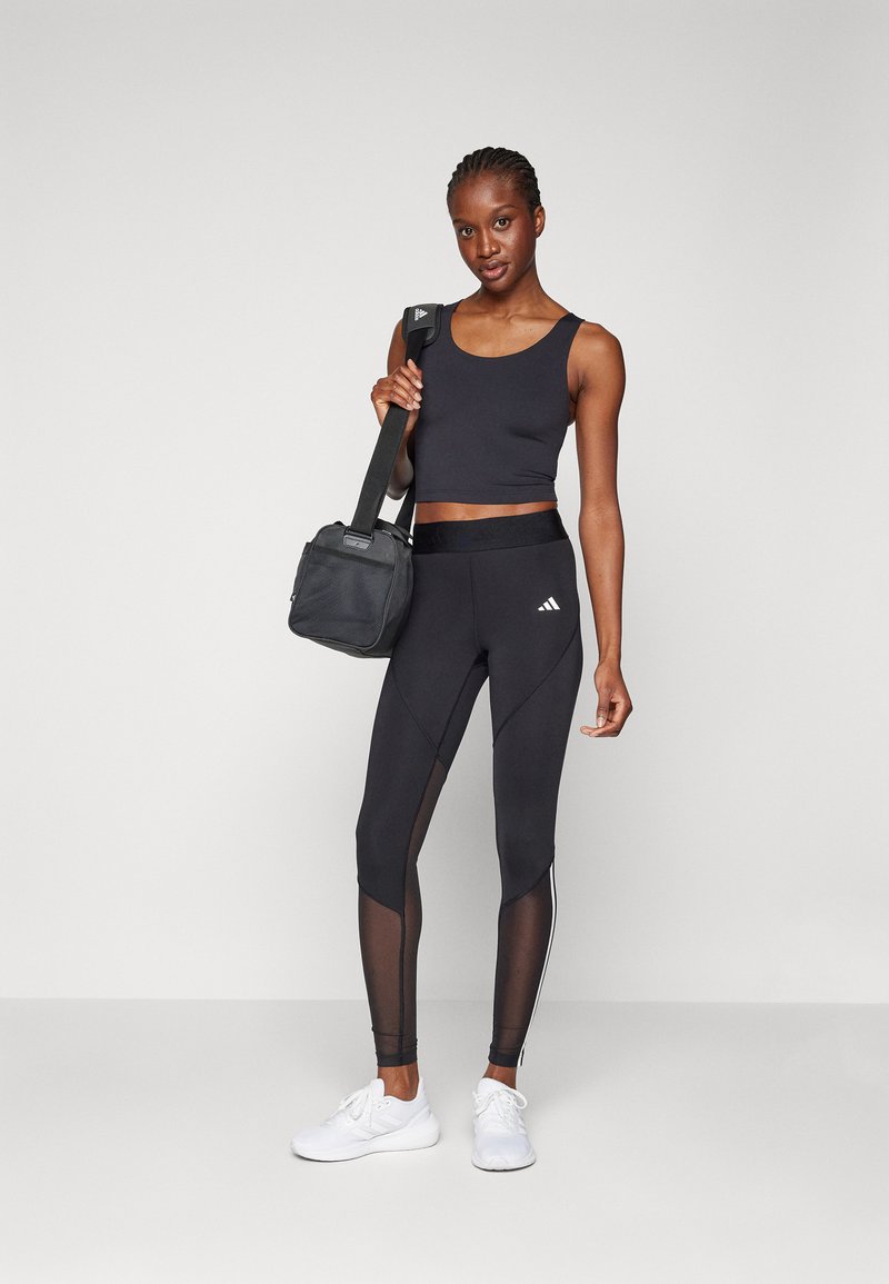 Haut de sport noir à bretelles larges, associé à un legging noir avec des panneaux en maille et des accents blancs. Tenant un petit sac noir.