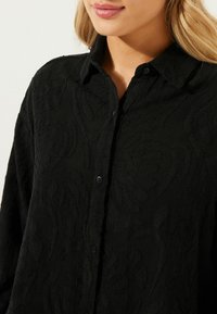Camicia nera con bottoni realizzata in tessuto testurizzato, caratterizzata da un motivo dettagliato. Collo classico e leggere sfilature lungo i bordi.