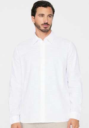 Mężczyzna o ciemnych włosach i brodzie, ubrany w białą teksturowaną koszulę z długim rękawem i guzikami, patrzący lekko w prawo, na jednolitym tle.