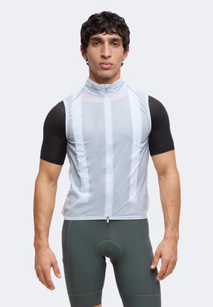 Homme portant un gilet de cyclisme sans manches bleu clair sur un t-shirt noir à manches courtes et un short de cyclisme rembourré vert foncé, debout face à l’avant.