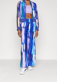 Persona che indossa un tuta tie-dye blu, viola e verde acqua con una giacca corta e pantaloni a gamba larga, scarpe da ginnastica bianche e un reggiseno sportivo lavanda.