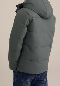 Grön puffjacka med huva, med kviltade sektioner och en slät, glänsande yta. Sammanvävd med blå denimjeans.