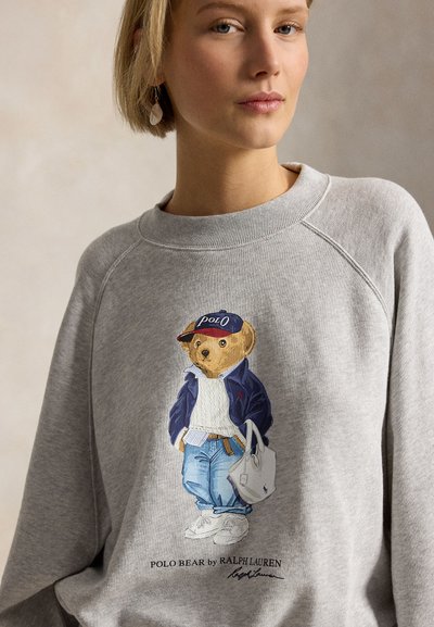 Γυναίκα που φορά γκρι φούτερ με γραφικό Polo Bear ντυμένο με τζιν, μπουφάν, αθλητικά παπούτσια και καπέλο, κρατώντας μια τσάντα tote.
