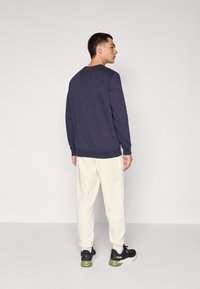 Sweatshirt bleu marine à manches longues, avec des poignets et un ourlet côtelés, associé à un pantalon de survêtement crème. Modèle portant des baskets noires avec des touches de vert.