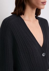 Schwarze Strickjacke mit tiefem V-Ausschnitt, strukturiertem Muster und Knöpfen vorne. Verfügt über gerippte und durchbrochene Details.