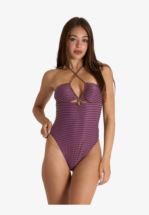 Donna che indossa un costume intero a righe viola e nere con spalline halter e un piccolo dettaglio a occhiello sul petto su sfondo bianco.