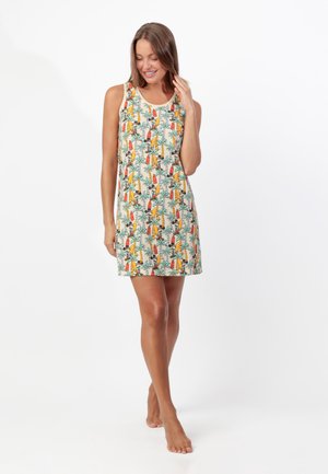 Mujer con vestido sin mangas de estampado tropical con palmeras y tablas de surf, de pie descalza con los ojos cerrados y sonriendo.