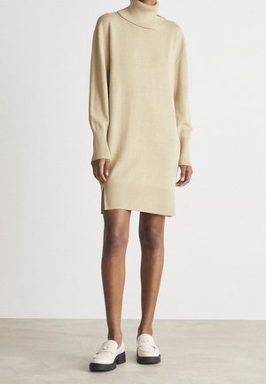 Beige strikket sweaterkjole med høj hals, lange ærmer og sideslids, kombineret med cremefarvede chunky loafers på en neutral baggrund.