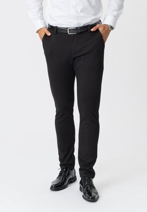 Schwarze Slim-Fit-Hosen aus einem glatten, dehnbaren Stoff, mit seitlichen Taschen und kombiniert mit schwarzen Dressschuhen.