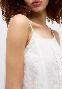 Γυναίκα με σγουρά μαλλιά φοράει λευκή αμάνικη μπλούζα με λεπτούς ιμάντες και δαντελένιες λεπτομέρειες, εμφανιζόμενη από τον ώμο μέχρι τα χείλη σε απλό φόντο.