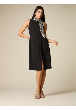 Frau in ärmellosem schwarzem Midikleid mit vorderem Schlitz, an den Hals gebundener Schal mit Tiermuster, passende Sandaletten mit hohem Absatz im Tiermuster und goldene Ohrringe.