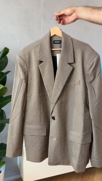 Brązowa i czarna kraciasta marynarka na drewnianym wieszaku. Posiada klapowy kołnierz, zapinana jest na guziki z przodu i ma kieszenie z patkami.