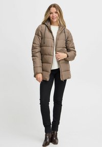 Doudoune marron avec capuche, design matelassé, cordon de serrage réglable et fermeture éclair, associée à un jean skinny noir et des bottines marron.
