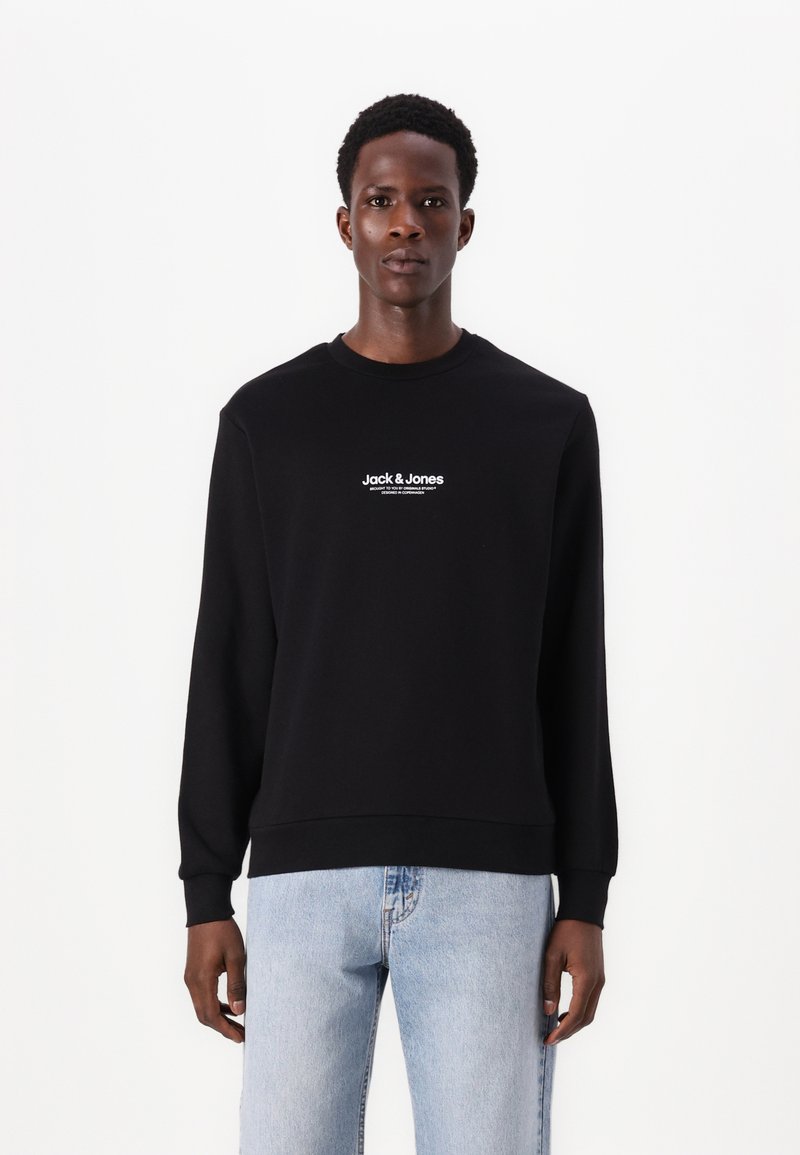 Mann in einem schwarzen Langarm-Sweatshirt mit dem Text "Jack & Jones" und hellblauen Jeans, der vor einem weißen Hintergrund steht.