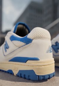 White and blue athletic shoe with mesh upper and textured sole, photographed outdoors on concrete with a blurred building in the background.