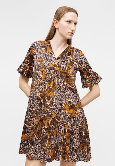 Γυναίκα που φοράει φόρεμα με κοντά μανίκια και V-λαιμόκοψη με πορτοκαλί, ναυτικό μπλε και μπεζ λουλουδένια σχέδια, στέκεται με τα χέρια πίσω από την πλάτη, κοιτάζοντας δεξιά.