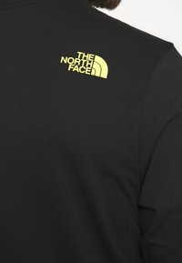 Nahaufnahme eines schwarzen North Face Shirts, das das gelbe Logo auf der oberen linken Seite des Rückens zeigt.