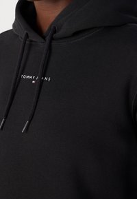 Gros plan d'un sweat à capuche noir avec cordons, avec le texte blanc "Tommy Jeans" et un petit logo de drapeau rouge, blanc et bleu sur la poitrine.