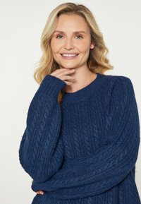 Maglione a coste di un blu navy con maniche lunghe e ampie; texture morbida; scollo rotondo; indossato da una persona con le mani che toccano il mento.