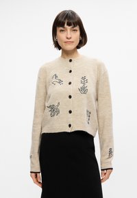 Μπεζ κάρντιγκαν από μαλλί με cropped σχέδιο, που διαθέτει μαύρα κεντημένα μοτίβα με άλογο, φυτό και γεωμετρικά στοιχεία; κλείσιμο με κουμπί.