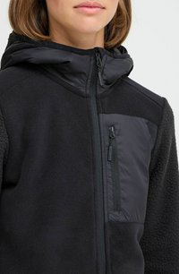 Veste hybride noire avec un corps en polaire confortable et une capuche en tissu lisse et élégant. Elle comprend une poche zippée sur la poitrine et une fermeture éclair.