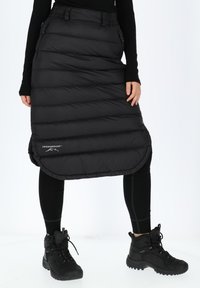 Black, puffy, insulated skirt with a curved hem, featuring a waistband and side pockets, paired with black leggings and sturdy boots.